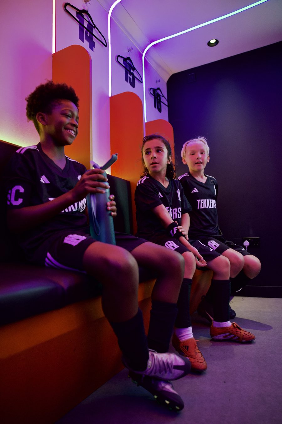 Three young players in Tekkers kit waiting on a changing-room bench